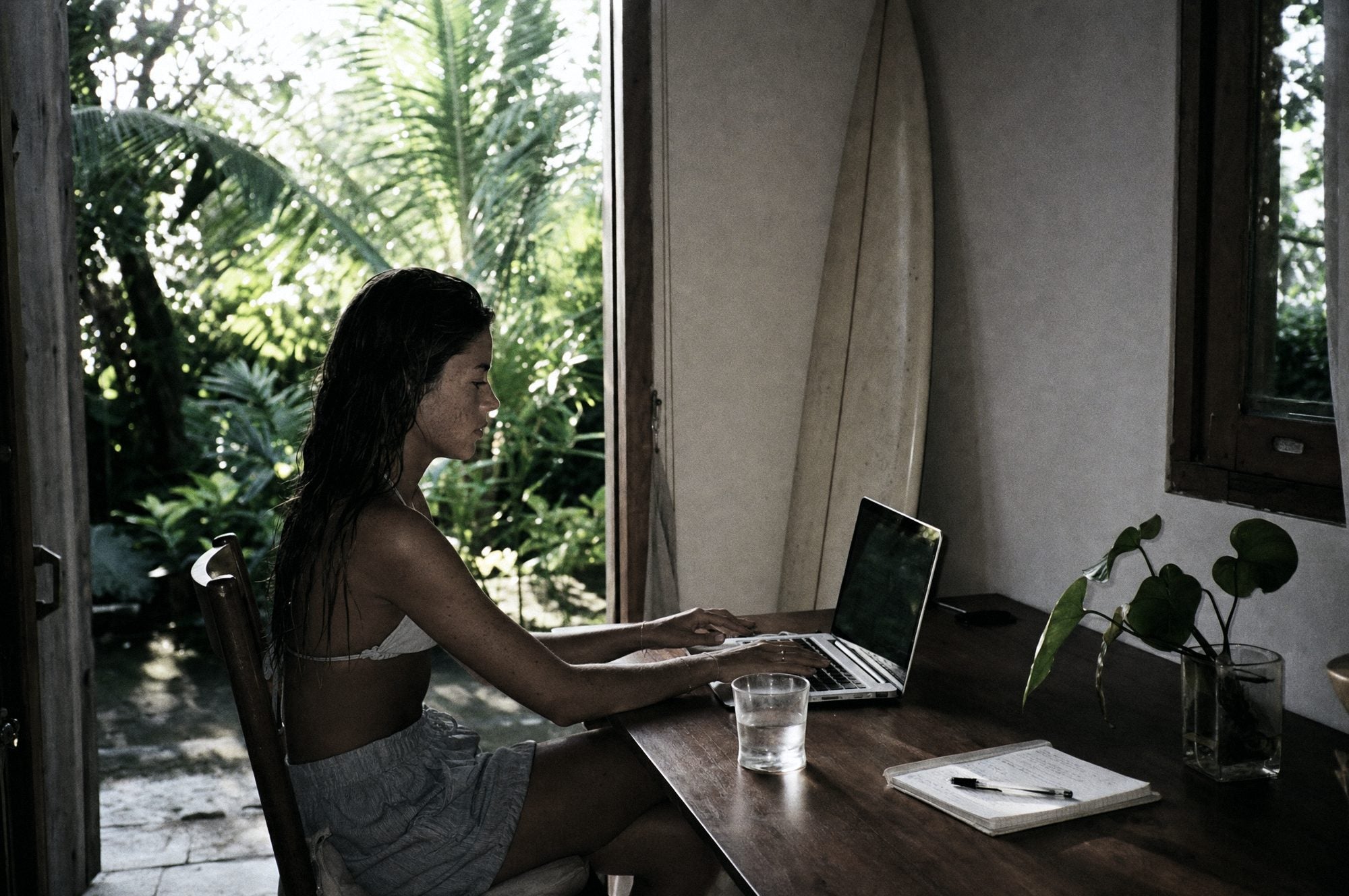 Back from a morning surf, wet hair, sitting down to work with a glass of water, a surfboard against the wall